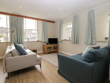 A living room with two sofas a wooden TV stand and windows with patterned curtains at The Old Haberdashery in Swanage