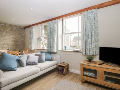 A living room with a sofa and cushions next to three windows with patterned curtains and a wooden TV stand with a plant at The Old Haberdashery in Swanage