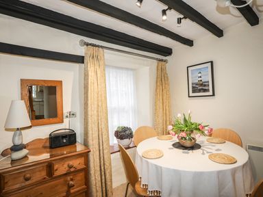 A dining area with a round table set with placemats and flowers next to a window with curtains and a wooden chest of drawers with a lamp and radio at Bwthyn Dwyfor in Beaumaris