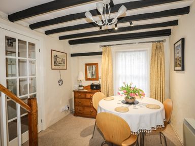 A dining area with a round table set with placemats and flowers a chest of drawers with a lamp and radio and a window with curtains at Bwthyn Dwyfor in Beaumaris