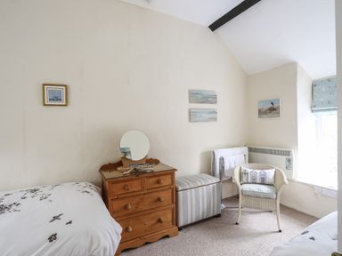 A bedroom with two single beds a wooden dresser with a round mirror a striped storage bench a wicker chair and paintings on the wall at Bwthyn Dwyfor in Beaumaris