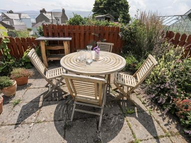 A round wooden table with four wooden chairs on a stone patio surrounded by plants and a wooden fence at Bwthyn Dwyfor in Beaumaris