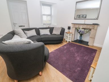 A living room with a curved gray sofa a purple rug and flowers in a fireplace at Rose Bank Cottage in St Just