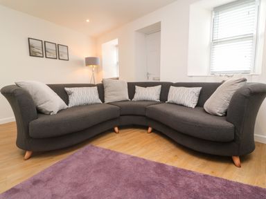 A dark curved sofa with cushions on a wooden floor near a window and lamp at Rose Bank Cottage in St Just