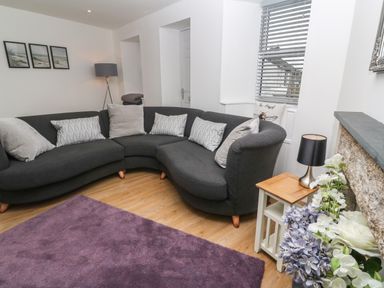 A living room with a curved dark grey sofa with cushions a purple rug a floor lamp and flowers at Rose Bank Cottage in St Just