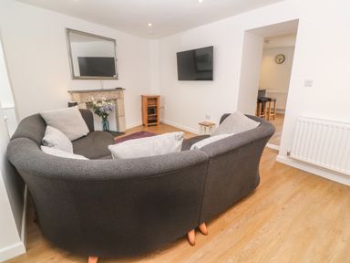 A living room with a curved dark sofa and cushions a wall-mounted TV a small wooden cabinet a mirror above a stone fireplace and a vase with flowers at Rose Bank Cottage in St Just