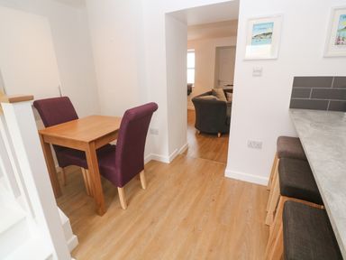 A small dining area with a wooden table and two purple chairs next to a kitchen counter with two stools and an open doorway leading to a living room at Rose Bank Cottage in St Just