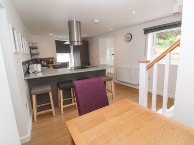 A kitchen with a breakfast bar and stools a wooden dining table with a purple chair a staircase and a window at Rose Bank Cottage in St Just