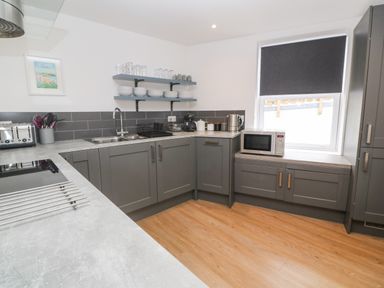 A kitchen with grey cabinets a microwave on the counter shelves with dishes and glassware and wooden floor at Rose Bank Cottage in St Just