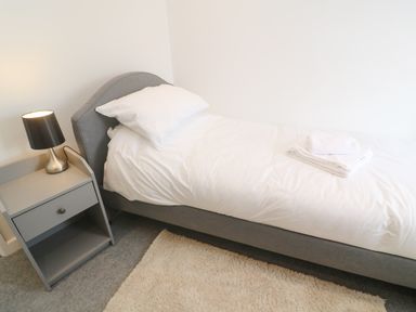 A single bed with white bedding and folded towels next to a gray nightstand with a lamp on a carpeted floor at Rose Bank Cottage in St Just