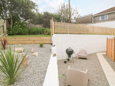 An outdoor patio area with gravel ground a barbecue grill two chairs wooden fences and plants at Rose Bank Cottage in St Just