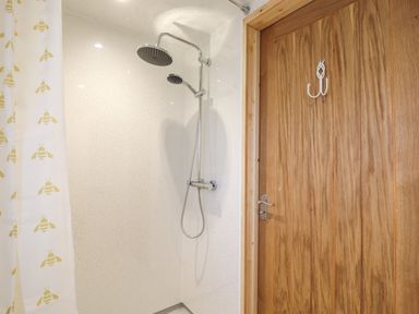 A shower area with a dual shower head and a bee patterned curtain next to a wooden door with a white hook at Buttercup Lodge in Theberton near Leiston