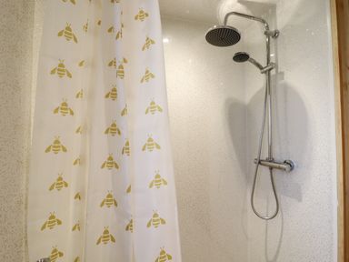 A shower area with a chrome showerhead and a white shower curtain with yellow bee patterns at Buttercup Lodge in Theberton near Leiston