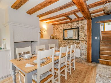 A dining room with a wooden table set for six white chairs a stone wall exposed wooden beams and stairs at Fox Den Cottage Castle Combe