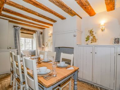 A dining room with a wooden table set for six with white chairs a fireplace and exposed wooden ceiling beams at Fox Den Cottage in Castle Combe