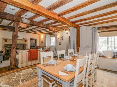 A dining area with a wooden table set with dishes and chairs next to a living room with sofas and a fireplace at Fox Den Cottage in Castle Combe
