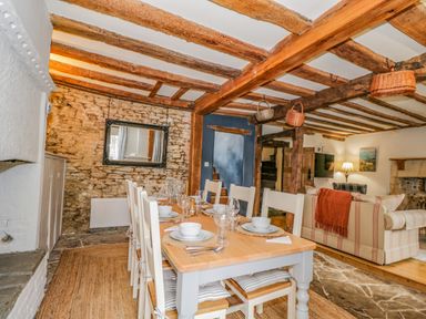 A dining area with a wooden table set for six chairs under wooden beams adjacent to a living room with sofas at Fox Den Cottage in Castle Combe