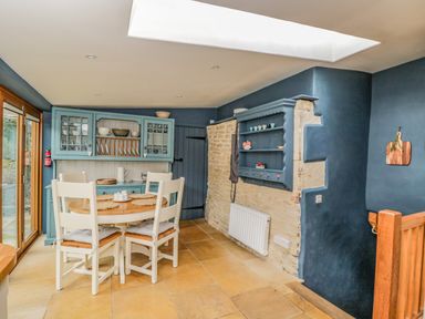 A dining area with a round wooden table and four white chairs near a blue cabinet and stone wall at Fox Den Cottage in Castle Combe