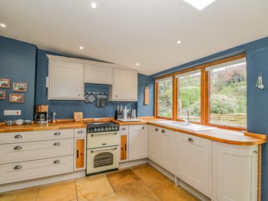 A kitchen with cream cabinets wooden countertops a stove a sink under large windows and blue walls at Fox Den Cottage in Castle Combe