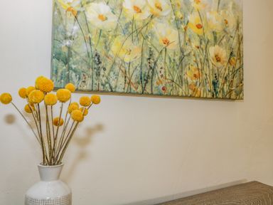 A white vase with yellow ball flowers on a wooden surface under a painting of yellow flowers at Fox Den Cottage in Castle Combe