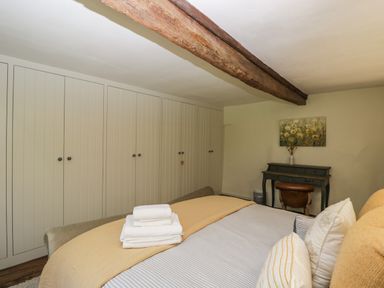A bedroom with a bed with pillows and folded towels and a wall of wardrobes and a desk with a chair and a painting at Fox Den Cottage in Castle Combe