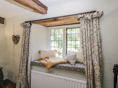 A window seat with cushions and a blanket by a window with floral curtains and wooden beams at Fox Den Cottage Castle Combe