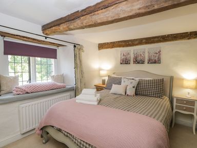 A bedroom with a bed with checkered bedding and pink blanket two bedside tables with lamps a window seat with cushions and a pink knitted throw at Fox Den Cottage Castle Combe