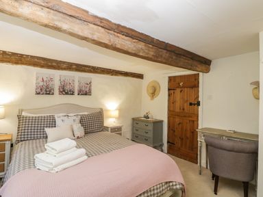 A bedroom with a bed with checked bedding two nightstands a wooden door a dresser a desk and a chair with wooden ceiling beams at Fox Den Cottage in Castle Combe