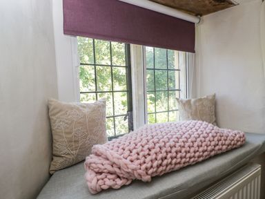 A window seat with a gray cushion two pillows and a folded pink knitted blanket at Fox Den Cottage in Castle Combe