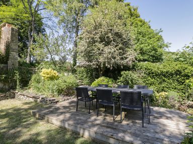 An outdoor seating area with a black table and six chairs on a wooden deck surrounded by trees and bushes at Fox Den Cottage in Castle Combe