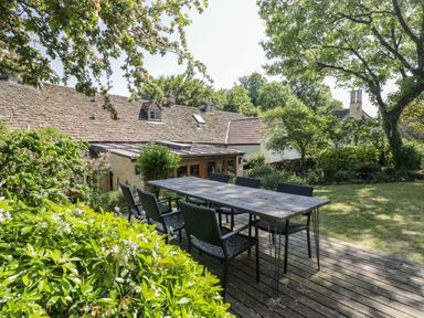An outdoor wooden deck with a dining table and chairs in a garden with trees and shrubs at Fox Den Cottage in Castle Combe