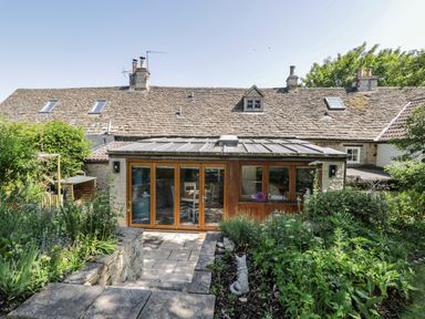 A garden with a variety of plants and a stone path leading to a house with a tiled roof and glass doors at Fox Den Cottage in Castle Combe