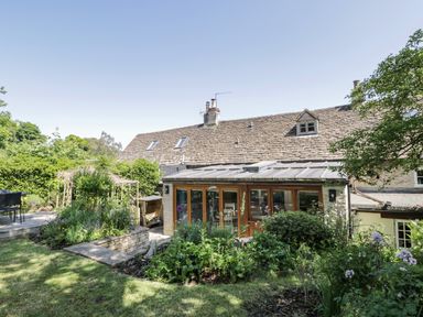 Garden with plants and a lawn in front of a cottage with a tiled roof and a glass enclosed porch at Fox Den Cottage in Castle Combe