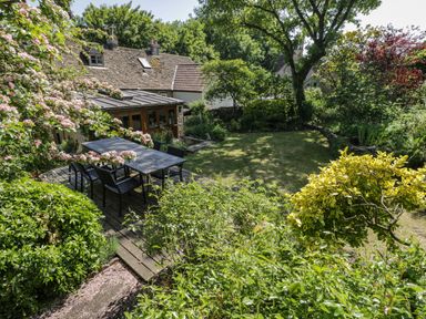 An outdoor wooden dining table with chairs on a wooden deck surrounded by trees and bushes at Fox Den Cottage in Castle Combe