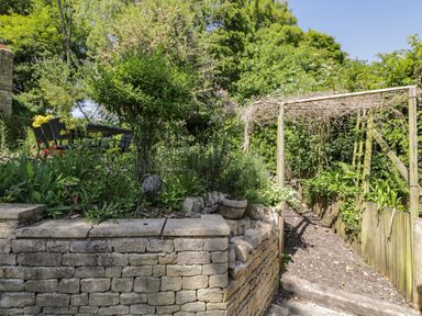 A garden with stone retaining wall patio table and chairs and wooden pergola at Fox Den Cottage in Castle Combe