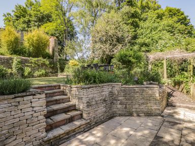 A garden with stone steps and retaining walls leading to a patio and outdoor dining area at Fox Den Cottage in Castle Combe