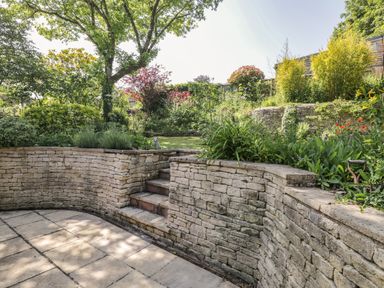 A garden with stone retaining walls and stairs leading to a lawn at Fox Den Cottage in Castle Combe