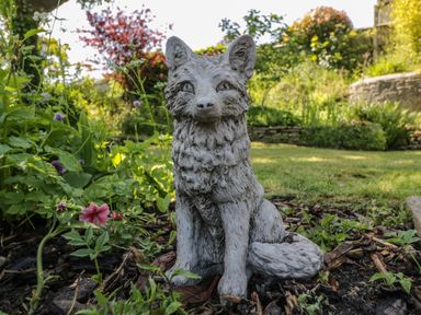 A garden with a stone fox statue surrounded by plants and flowers at Fox Den Cottage in Castle Combe