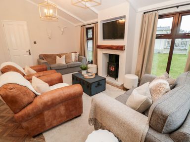 A living room with two brown armchairs two grey sofas a television above a fireplace and windows with beige curtains at The Treeline Swarland near Felton