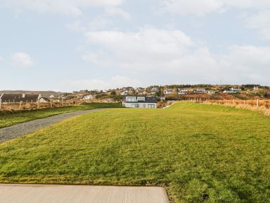 A grassy field with a narrow gravel path leading to a house and a village on a hillside in the background at The Beachhouse in Meenlaragh near Gortahork County Donegal