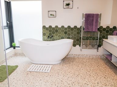 A bathroom with a freestanding white bathtub green hexagonal tiles on the wall a towel rack with purple towels and a window with a small plant at The Beachhouse in Meenlaragh near Gortahork County Donegal