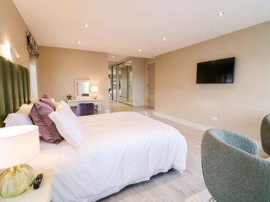 A bedroom with a bed a bedside lamp a desk with a chair and mirror a wall mounted TV and gray chairs at The Beachhouse in Meenlaragh near Gortahork County Donegal