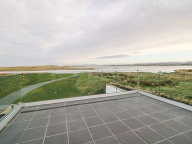 A balcony with glass railing overlooking a grassy landscape and a body of water at The Beachhouse in Meenlaragh near Gortahork County Donegal