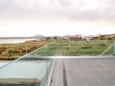 A balcony with glass railings overlooking grassy fields houses a coastline and mountains at The Beachhouse in Meenlaragh near Gortahork County Donegal