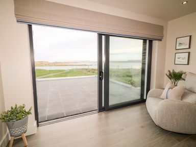 A room with a sliding glass door opening to a patio and views of water with a gray armchair and potted plants at The Beachhouse in Meenlaragh near Gortahork County Donegal