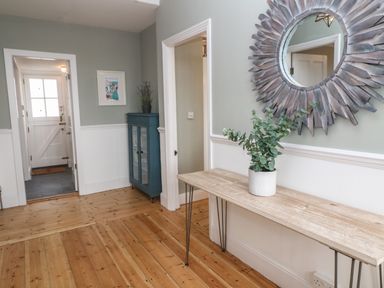 A hallway with a wooden floor a wooden console table with a plant a round sunburst mirror a blue cabinet and a white door at Belmont in Newlyn