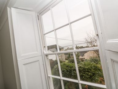 A window with white shutters overlooking bushes houses and a body of water at Belmont in Newlyn