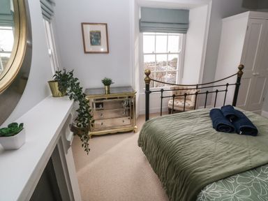 A bedroom with a bed covered with a green quilt and blue towels a mirrored dresser plants a beige chair and a white wardrobe at Belmont in Newlyn