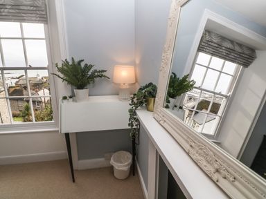 A small white table with plants and a lamp next to a window with a view of rooftops and a large decorative mirror reflecting the window at Belmont in Newlyn