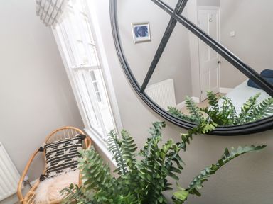 A room corner with a window wicker chair with cushions green plant and a round wall mirror at Belmont in Newlyn